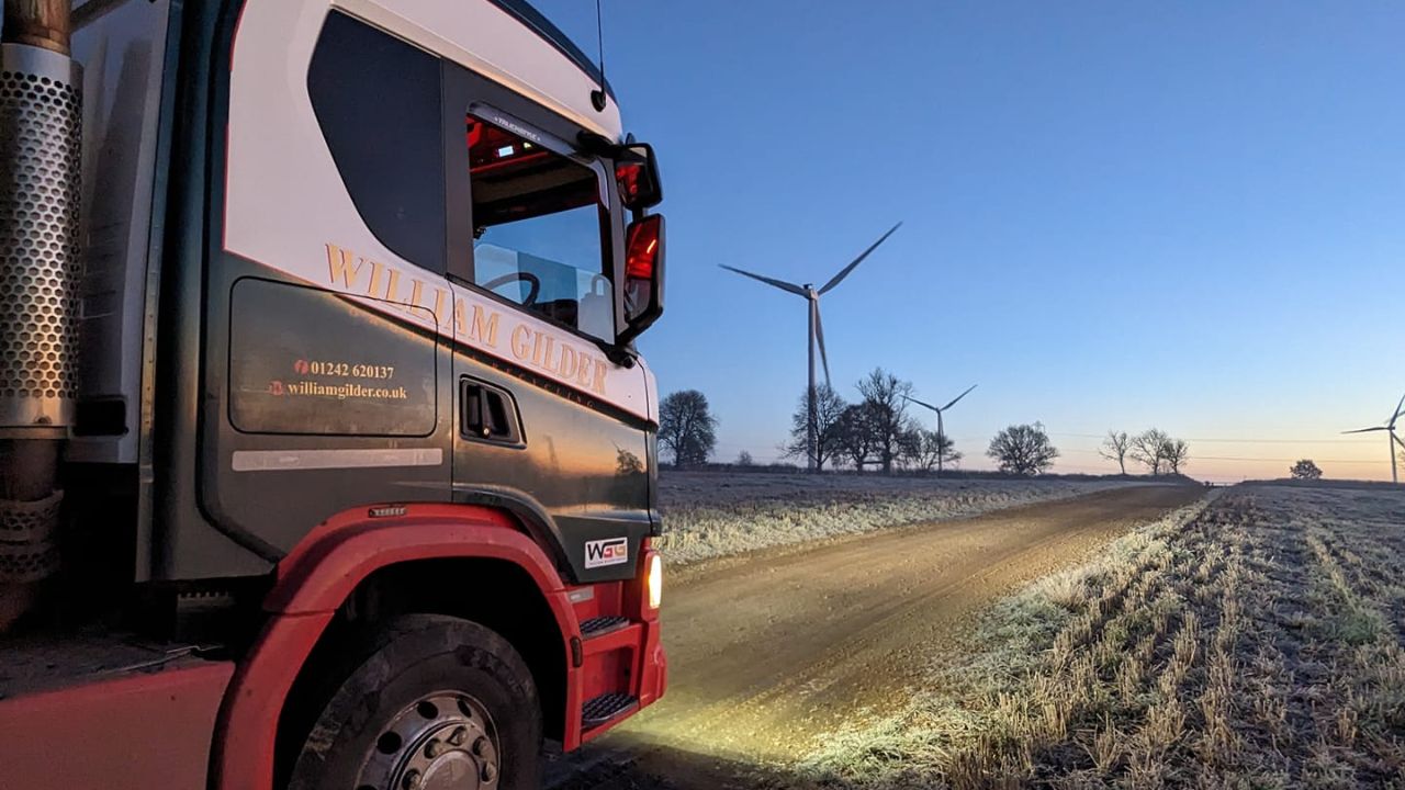 A lorry on a snow covered road with a wind turbine in the distance